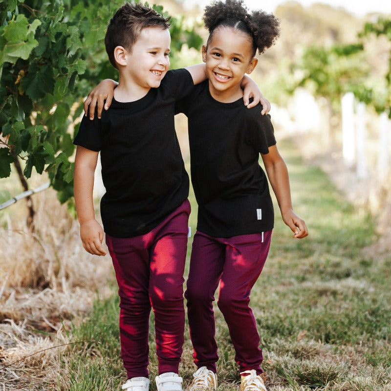 boy and girl standing in between the winery vines with their arms around each others shoulders. Both are smiling and wearing Frankie & Roy maroon rye pants and black T-Shirt
